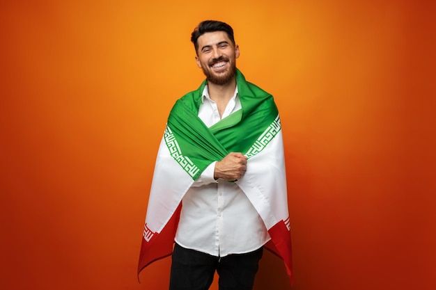 Porträt eines jungen, lässigen Mannes, der die Flagge des Iran vor orangefarbenem Hintergrund hält