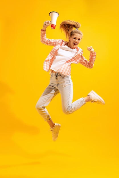 Porträt eines jungen Mädchens, das springend mit Megaphon posiert, isoliert über gelbem Studiohintergrund