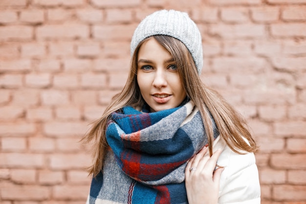 Porträt eines jungen Mädchens in einer Strickmütze, einer weißen Jacke und einem hellen Schal nahe einer Mauer