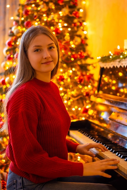 Porträt eines jungen Mädchens in leuchtend rotem Pullover beim Spielen des Klaviers geschmückter Weihnachtsbaum im Hintergrund warmes Licht in der festlichen Atmosphäre des Raumes