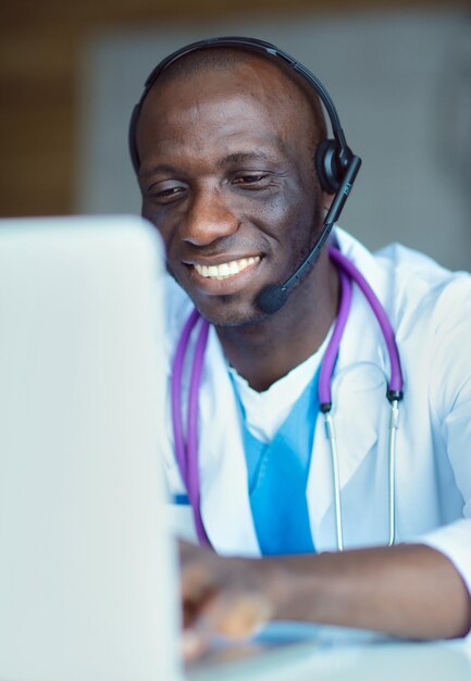 Porträt eines jungen männlichen Arztes, der ein Headset trägt, während er den Computer am Schreibtisch in der Klinik benutzt Doktor