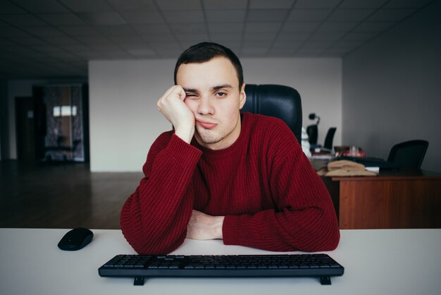 Porträt eines jungen Mannes, der mit dem Computer müde ist. Vor dem Hintergrund von Büroräumen. Die Situation im Büro