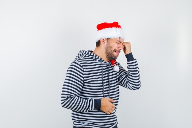 Porträt eines jungen Mannes, der sich das Auge reibt, während er in Hoodie, Weihnachtsmütze und deprimiert weint