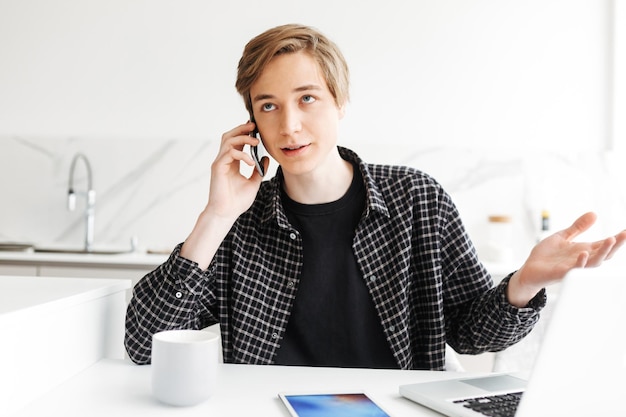 Porträt eines jungen Mannes, der sitzt und etwas bespricht, während er zu Hause auf seinem Handy in der Küche spricht