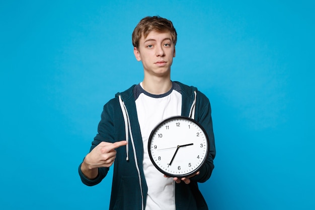 Porträt eines jungen Mannes in Freizeitkleidung, der mit dem Zeigefinger auf die runde Uhr zeigt, die auf der blauen Wand isoliert ist. Die Zeit wird knapp. Menschen aufrichtige Emotionen Lifestyle-Konzept.