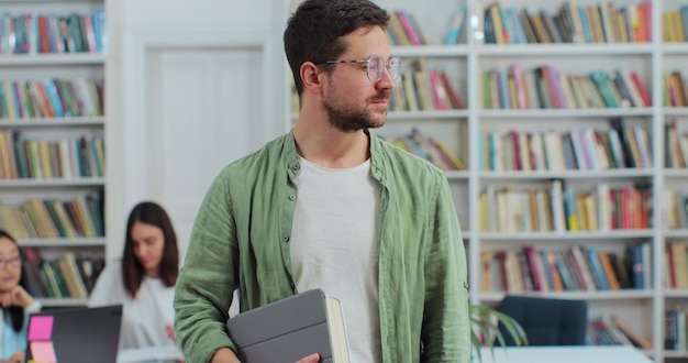 Porträt eines jungen Mannes mit Brille, der ein digitales Tablet im Bücherregal der Bibliothek hält und in die Kamera blickt Hochschulbildungskonzept