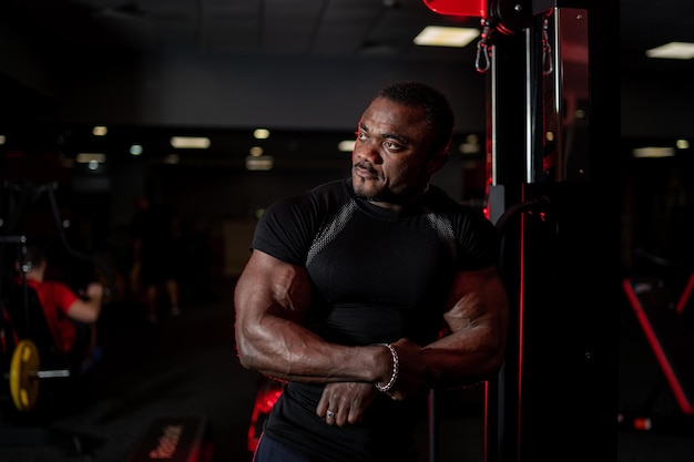 Porträt eines jungen muskulösen Afroamerikaners in Sportkleidung, der die Seite betrachtet, während er über dem Hintergrund des Fitnessstudios posiert. Sport, Training, Bodybuilding-Konzept.
