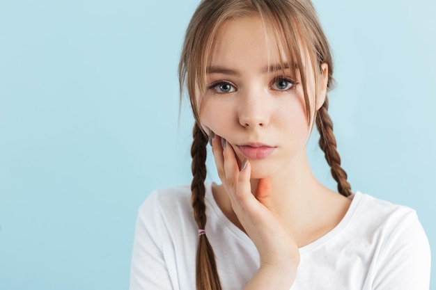 Porträt eines jungen nachdenklichen Mädchens mit zwei Zöpfen im weißen T-Shirt, das die Hand an der Wange hält, während es nachdenklich in die Kamera über blauem Hintergrund schaut