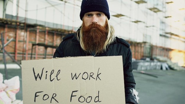 Porträt eines jungen Obdachlosen mit Pappe, der in die Kamera schaut und für Essen arbeiten will