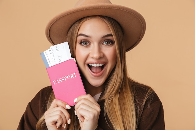 Porträt eines jungen optimistischen Mädchens mit Hut, das Reisepass und Reisetickets isoliert auf Beige hält