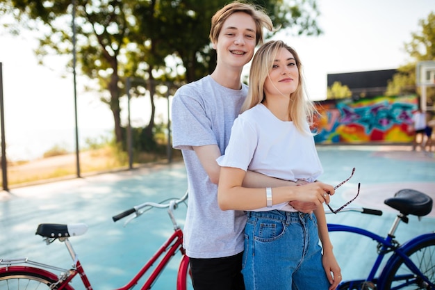 Porträt eines jungen Paares, das zusammensteht und glücklich mit Fahrrädern daneben in die Kamera schaut Fröhlicher Junge, der mit hübschem Mädchen steht und sie umarmt, während er Zeit auf dem Basketballplatz im Park verbringt