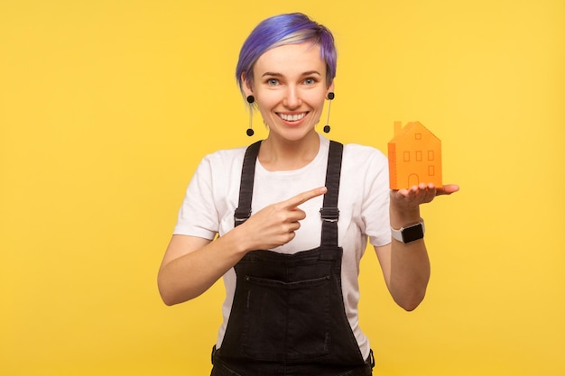 Porträt eines jungen, positiven, modischen Hipster-Mädchens mit violetten kurzen Haaren in Denim-Overalls, das auf ein orangefarbenes Papierhaus auf der Handfläche zeigt und lächelt. Studioaufnahme im Innenbereich isoliert auf gelbem Hintergrund