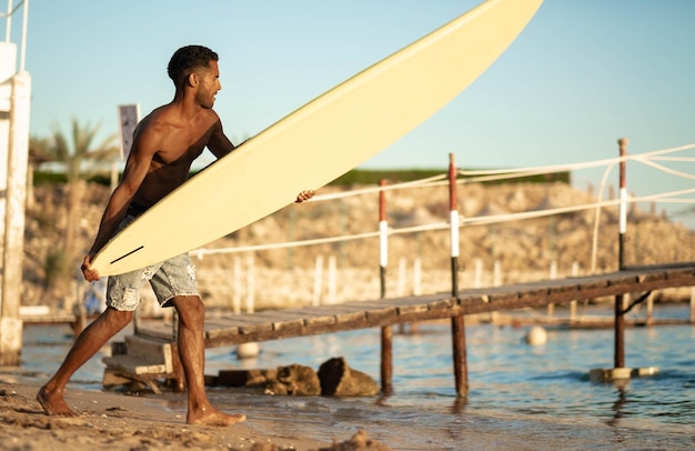 Porträt eines jungen professionellen schwarzen Surfers, der sein Surfbrett im Arm hält, steht am Strand bei Sonnenuntergang, Sonne hinter der Schulter, Sporthobby, entspannter Lebensstil