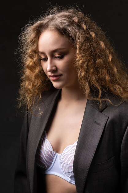 Porträt eines jungen, rothaarigen, lockigen, sexy Mädchens in einer Jacke mit langen Haaren im Studio. Zeigt ihre kurvenreichen Brüste in einem BH