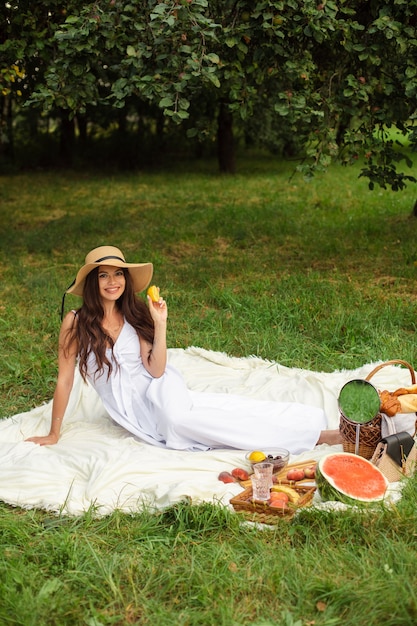 Porträt eines jungen schönen Mädchens mit sogar weißen Zähnen, einem schönen Lächeln in einem Strohhut und einem langen weißen Kleid machen ein Picknick im Garten.