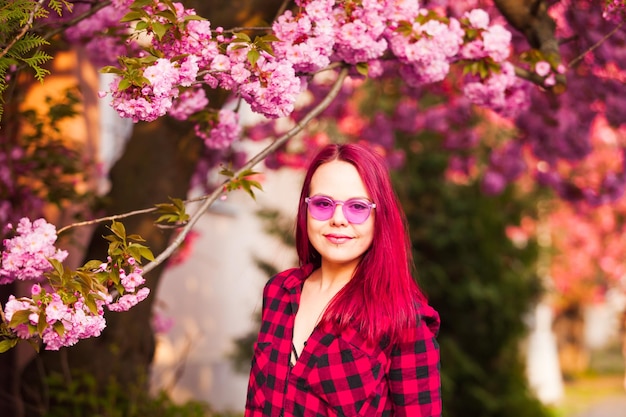 Porträt eines jungen, schönen Modells mit lebendigem Haar, das eine rosafarbene Sonnenbrille trägt und in die Kamera schaut, die in der Nähe eines blühenden Sakura-Baums posiert Brillante Haarfarben und Sakura-Blüte