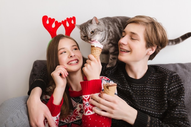 Porträt eines jungen schönen Paares, das zu Hause auf dem Sofa sitzt, mit Eis in den Händen und einer hübschen Katze in der Nähe, die Eis versucht