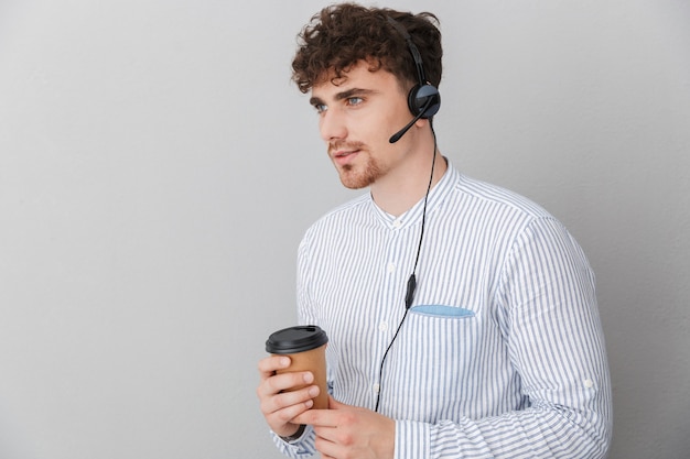 Porträt eines jungen, schönen Telefonisten, der ein Mikrofon-Headset trägt und eine Kaffeetasse hält, während er mit dem Kunden spricht, isoliert über grauer Wand