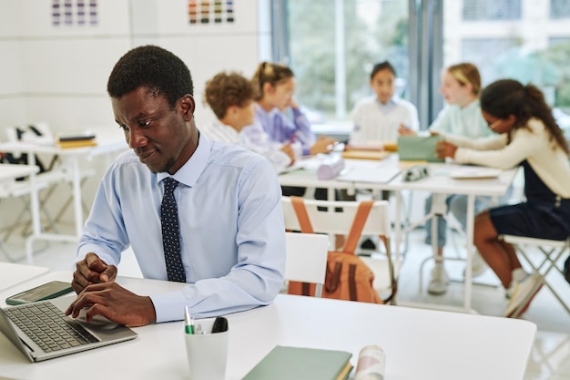 Porträt eines jungen schwarzen Lehrers mit Laptop im Klassenzimmer mit einer Gruppe von Kindern im Hintergrund