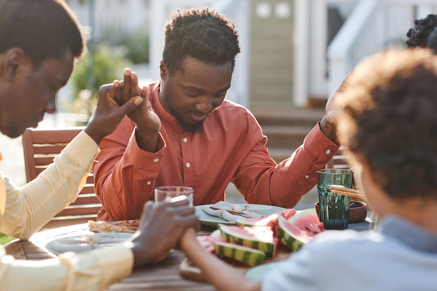 Porträt eines jungen schwarzen Mannes, der während des Familientreffens am Tisch im Freien Anmut sagt und sich an den Händen hält