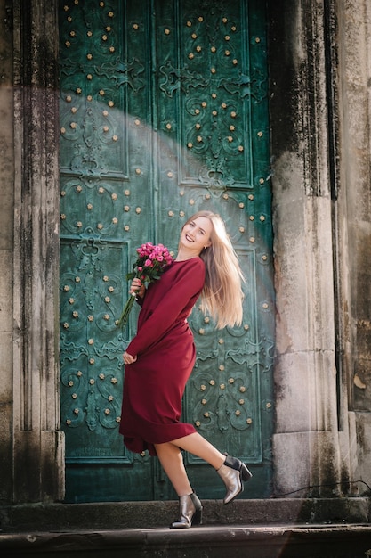 Porträt eines jungen, stilvollen, schönen Mädchens in einem roten Kleid mit Blumenstrauß, das auf der Straße läuft und lächelt, genießt ihre Wochenenden. Sommer, sonnig, trendy, Lifestyle.