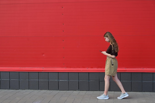 Porträt eines jungen Studenten, der eine Nachricht auf einem Handy liest, ein stilvolles, hübsches Mädchen, das auf der Straße unterwegs mit einem Handy plaudert, soziale Störung, Technologie, Krankheitsabhängigkeit