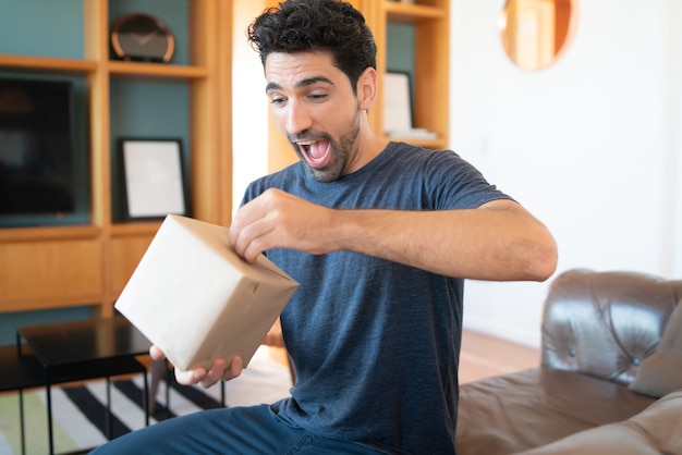 Porträt eines jungen überraschten Mannes, der eine Geschenkbox öffnet, während er zu Hause auf der Couch sitzt.