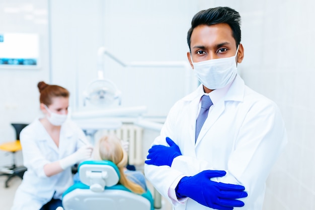 Porträt eines jungen überzeugten asiatischen männlichen Zahnarztes in der Klinik vor dem wfemale Zahnarzt, der Zähne zum Patienten überprüft. Zahnklinik-Konzept