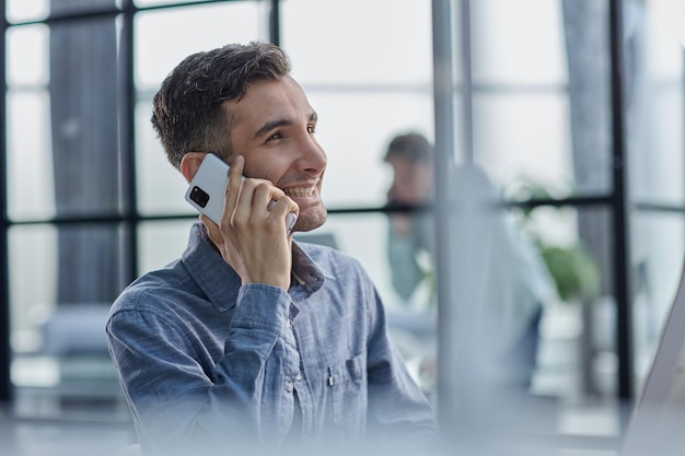 Porträt eines jungen Unternehmers in einem beiläufigen Büro, der einen Telefonanruf macht, während er mit einem Laptop arbeitet