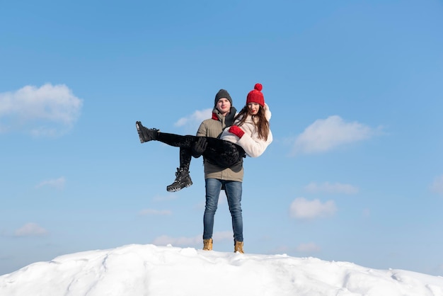 Porträt eines jungen verliebten Paares auf einem schneebedeckten Hügel vor blauem Himmel. Guy hält das Mädchen in seinen Armen.