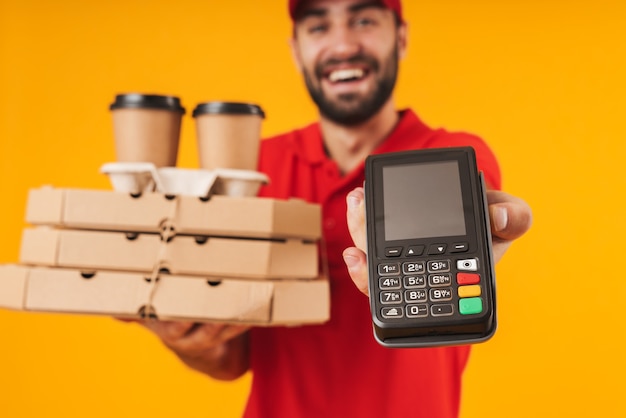 Porträt eines kaukasischen Lieferboten in roter Uniform mit Pizzakartons und Zahlungsterminal isoliert über Gelb