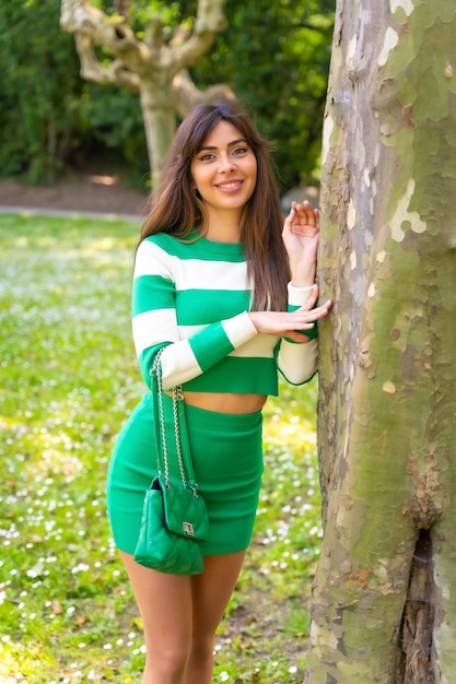 Porträt eines kaukasischen Mädchens in der Natur in einem grün-weißen Pullover und mit einem Taschenmode-Modell, das neben einem Baum posiert