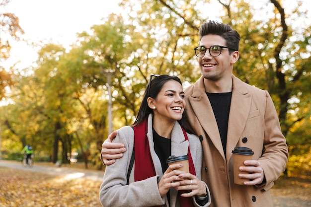 Porträt eines kaukasischen Paares, Mann und Frau der 20er Jahre, die Kaffee zum Mitnehmen aus Pappbechern beim Gehen im Herbstpark trinken