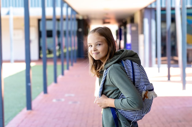 Porträt eines kaukasischen Schulmädchens mit Schultasche, das auf dem Schulflur zur Kamera lächelt