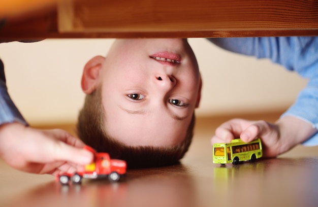 Porträt eines Kindes eines Vorschuljungen kopfüber mit einem Spielzeugauto und einem Bus in den Händen. Blick vom Boden unter dem Bett.