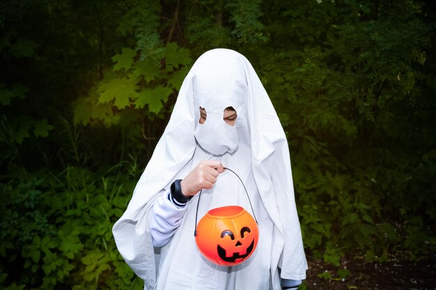 Porträt eines Kindes im Halloween-Geisterkostüm, das im dunklen Wald auf einen Kürbiskorb blickt