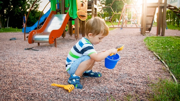 Porträt eines kleinen 3 Jahre alten Kleinkindjungen, der auf dem Spielplatz sitzt und Sand mit Spielzeugplastikschaufel gräbt