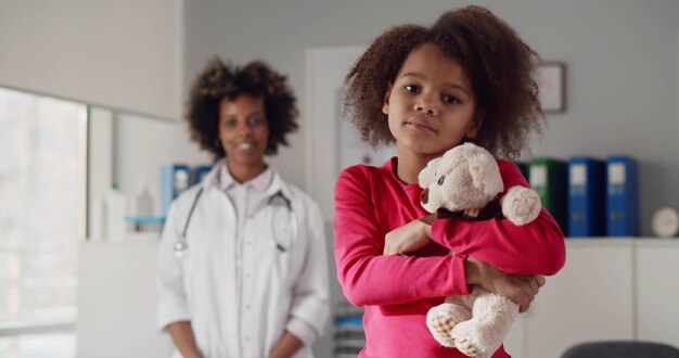 Foto porträt eines kleinen afroamerikanischen mädchens mit teddybär und einer jungen ärztin im hintergrund