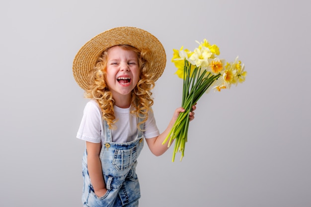 Porträt eines kleinen blonden Mädchens, das einen Blumenstrauß der Frühlingsblumen in einem Strohhut auf einem weißen Raum hält