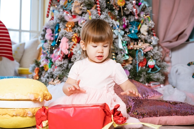 Porträt eines kleinen blonden Mädchens, das in der Nähe des Weihnachtsbaums spielt, das Kind trägt ein weißes Ts...