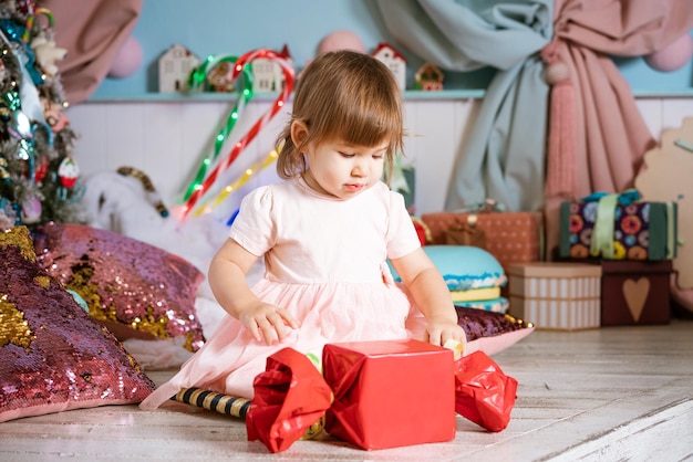 Porträt eines kleinen blonden Mädchens, das in der Nähe des Weihnachtsbaums spielt, das Kind trägt ein weißes Ts...