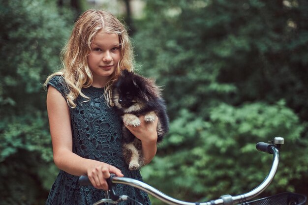 Porträt eines kleinen blonden Mädchens in einem lässigen Kleid, hält süßen Spitzhund. Fahren Sie mit dem Fahrrad in einem Park.