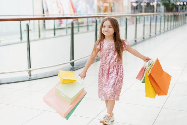 Porträt eines kleinen glücklichen Mädchens im Einkaufszentrum. Ein lächelndes lachendes Mädchen in einem rosa Kleid mit bunten Taschen in den Händen geht durch das Einkaufszentrum und schaut auf die Schaufenster