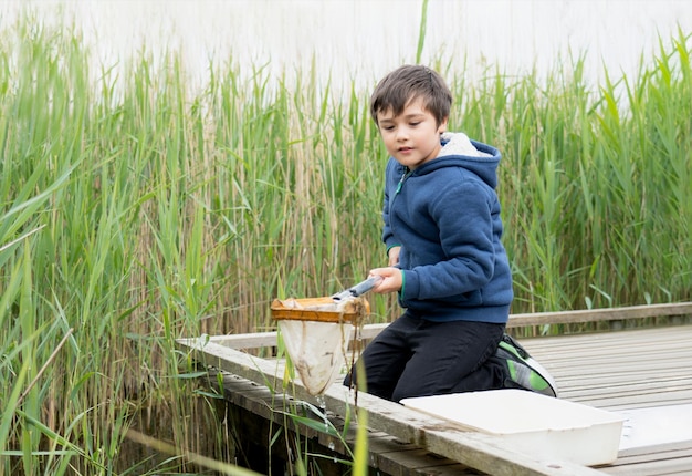 Porträt eines kleinen Jungen, der im Frühling Lebewesen im Teich mit einem Netz fängt. Kinderforscher und Lernen über die wilde Natur auf dem Land. Sommercamp im Freien für Kinder