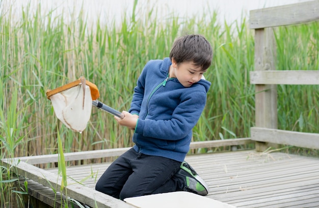 Porträt eines kleinen Jungen, der im Frühling mit einem Fischerkescher Lebewesen im Teich mit einem Netz fängt. Kinderentdecker und Lernen über die wilde Natur auf dem Land. Sommercamp im Freien für Kinder