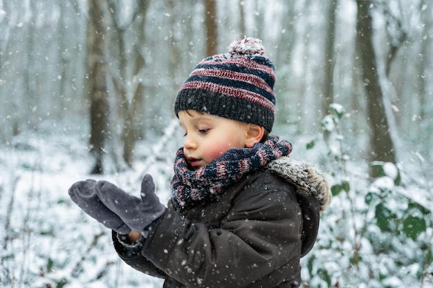 Porträt eines kleinen Jungen, der im Winter mit Schneeflocken in einem Park spielt, hautnah. Glückliches Kind genießt den ersten Schnee in einem Wald. Warten auf Weihnachten.