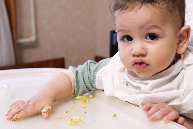 Porträt eines kleinen Jungen, der Nahrung isst. Baby mit einem Löffel im Fütterungsstuhl. Niedliches Baby, das seine erste Mahlzeit isst
