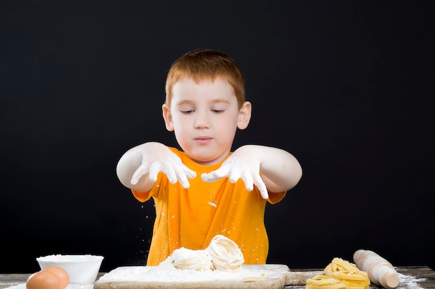 Porträt eines kleinen Jungen in der Küche, während er hilft, Essen zuzubereiten Ein Junge mit roten Haaren und schönen Gesichtszügen hilft einem Kind in der Küche, Nudeln zu kochen