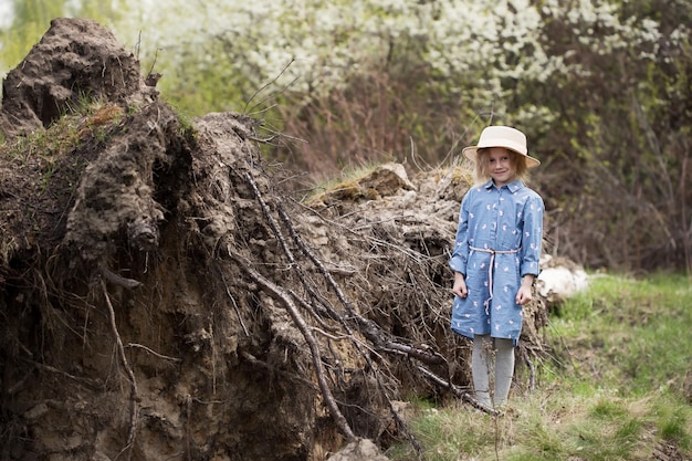 Porträt eines kleinen kaukasischen Mädchens, das neben einem umgestürzten Baum mit umgedrehten Wurzeln im Wald steht und in die Kamera schaut