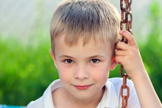 Porträt eines kleinen lächelnden Jungen mit goldenen blonden Strohhaaren im sonnigen Sommertag auf grünem unscharfem Hintergrund